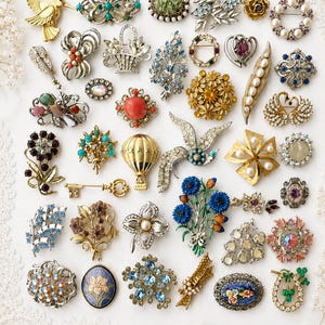 May include: A collection of assorted vintage-style brooches featuring various designs including birds, flowers, geometric shapes, and abstract patterns. The pins are crafted with colorful rhinestones, faux pearls, enamel, and metallic finishes in gold and silver tones.
