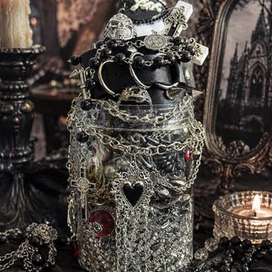 May include: A clear plastic jar filled with assorted silver-toned chains, black beads, and metal charms including a heart, a crucifix, and a skull ring. A black leather-like belt with metal rings and spikes is wrapped around the top of the jar.