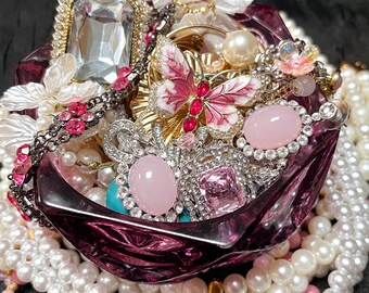 May include: A collection of jewelry pieces, including necklaces, rings, and brooches, displayed in a deep purple glass bowl. The jewelry features pearls, crystals, and gemstones in various colors, including pink, white, and gold. A butterfly brooch is a focal point.