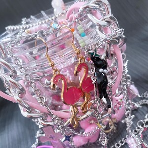 May include: A collection of pink and silver jewelry, including earrings with flamingo charms, bracelets, and necklaces. The earrings have gold hooks. The jewelry is arranged around a clear container with pink accents and silver chains.