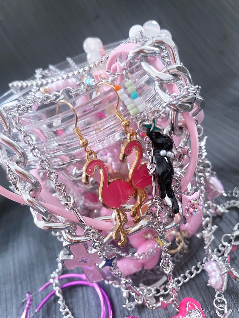 May include: A collection of pink and silver jewelry, including earrings with flamingo charms, bracelets, and necklaces. The earrings have gold hooks. The jewelry is arranged around a clear container with pink accents and silver chains.
