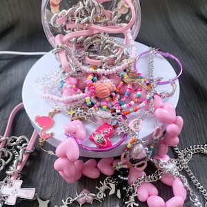 May include: A collection of pink and silver jewelry, including necklaces, bracelets, and earrings, spills from a clear container. The jewelry features heart, cross, and star charms, along with beaded and chain designs. A flamingo charm is also visible.