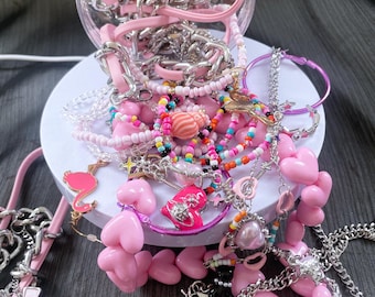 May include: A collection of pink and silver jewelry, including necklaces, bracelets, and earrings, spills from a clear container. The jewelry features heart, cross, and star charms, along with beaded and chain designs. A flamingo charm is also visible.