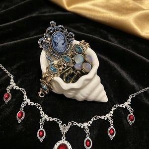 Op de afbeelding: Een zilverkleurige ketting met meerdere bungelende rode ovale cabochons en een wit keramieken schelpvormig schaaltje met een collectie sieraden, waaronder een blauwe cameebroche met kleine blauwe kristallen en diverse metalen armbanden.