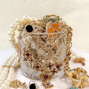 May include: A clear glass jar overflowing with various jewelry pieces. Gold necklaces, bracelets, and rings are mixed with pearl strands and a jeweled butterfly. A red and gold floral necklace is in the foreground, with a gold mermaid pendant.