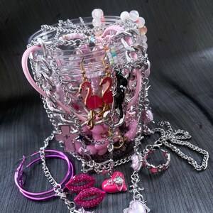 May include: A collection of pink and silver jewelry, including earrings, necklaces, and rings, spilling from a clear container. The earrings feature flamingo and cat designs. Other items include a lip-shaped pendant and a heart-shaped necklace.