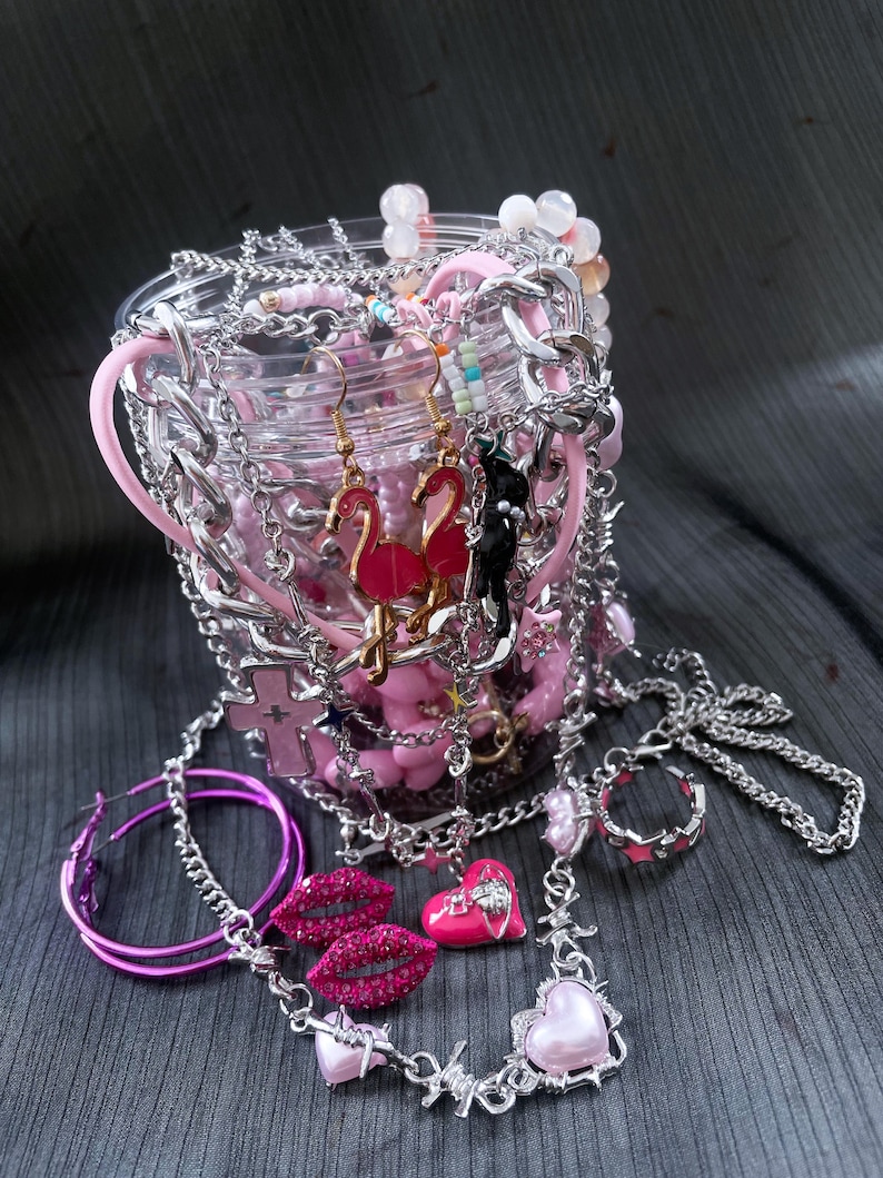 May include: A collection of pink and silver jewelry, including earrings, necklaces, and rings, spilling from a clear container. The earrings feature flamingo and cat designs. Other items include a lip-shaped pendant and a heart-shaped necklace.