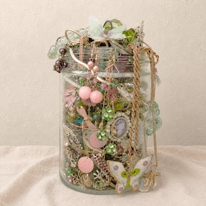 May include: A clear glass jar filled with a collection of assorted jewelry, including gold-tone chains, pink bead earrings, green crystal accents, and a prominent white butterfly-shaped pendant with green and pink details resting on the front.