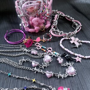 May include: A collection of jewelry including a clear plastic jar filled with pink beads and charms, silver-tone chain necklaces with pink heart pendants, barbed wire style chains, hoop earrings, and a beaded necklace with a pink cross charm.