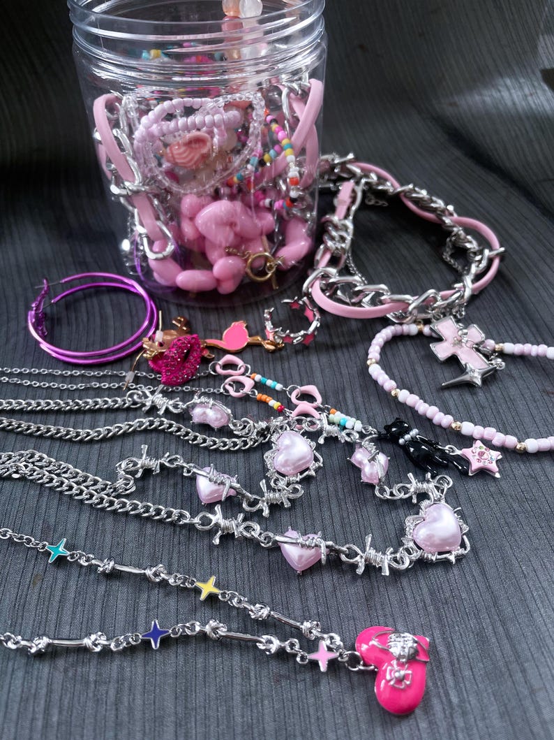 May include: A collection of jewelry including a clear plastic jar filled with pink beads and charms, silver-tone chain necklaces with pink heart pendants, barbed wire style chains, hoop earrings, and a beaded necklace with a pink cross charm.