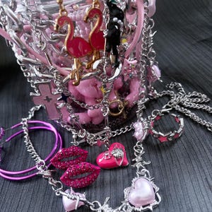 May include: A collection of pink and silver jewelry, including earrings with flamingo charms, a necklace with heart pendants, and a ring. The jewelry is displayed in a clear container with pink accents and chains.