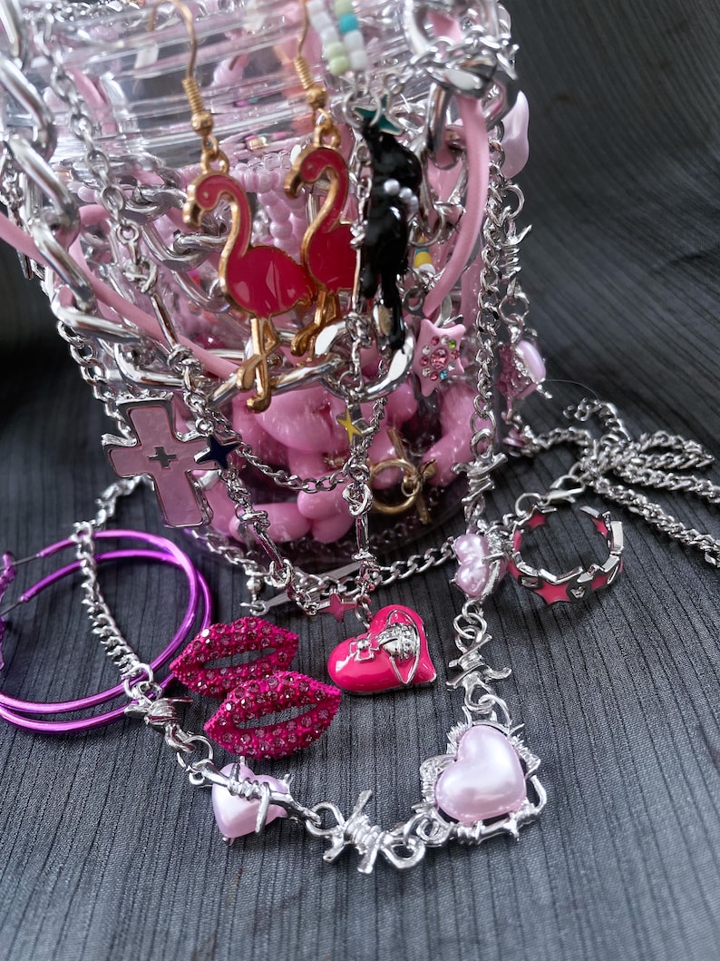 May include: A collection of pink and silver jewelry, including earrings with flamingo charms, a necklace with heart pendants, and a ring. The jewelry is displayed in a clear container with pink accents and chains.