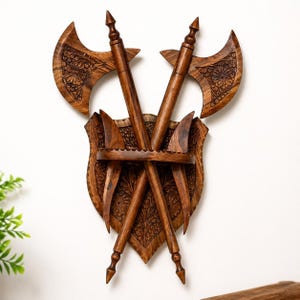 May include: Wooden wall decor featuring two crossed axes and a shield. The axes and shield are intricately carved with floral patterns. The axes have pointed tips and curved blades. The wood has a warm, brown tone, and the design is mounted on a white wall.
