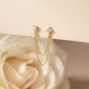 May include: A pair of gold-coloured double-chain earrings. One earring has a square-shaped clear stone, the other a cluster of smaller clear stones in a floral arrangement. The chains hang below the ear studs.