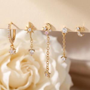 May include: A collection of gold-toned earrings featuring celestial designs. The set includes a hoop earring with a star charm, a starburst drop earring, a moon and star chain earring, a round stud earring with a chain and a crescent moon and star stud.
