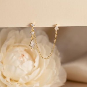 Könnte beinhalten: Ein goldfarbener Doppelohrring mit zarter Kette. Ein Ende hat einen kleinen, funkelnden, rautenförmigen Edelstein, das andere eine Ansammlung von drei kleineren Edelsteinen und einen hängenden, rautenförmigen Edelstein. Der Hintergrund ist weich und neutral.