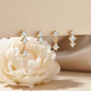 May include: Four pairs of gold-colored earrings with clear, sparkling cubic zirconia stones. The earrings have a three-stone design, with one pair featuring a dangling stone. The earrings are displayed against a soft, neutral background.