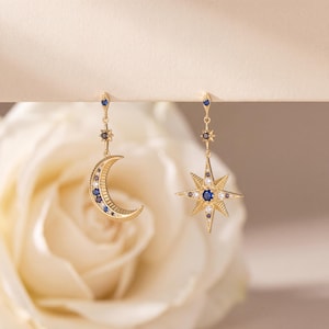 May include: A pair of gold-toned drop earrings featuring celestial motifs. One earring displays a crescent moon shape, while the other is a multi-pointed star. Both pieces are accented with small, embedded blue and clear stones throughout.
