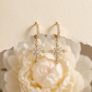 May include: A pair of gold hoop earrings featuring dangling cross pendants. Each cross is encrusted with clear baguette and round-cut stones, creating a radiant, symmetrical design. The hoops are thin and circular, connecting to the cross charms.