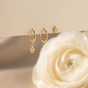 May include: A set of three gold-colored earrings. Two hoop earrings and a stud earring are visible. One hoop earring has a diamond-shaped charm with a small clear stone. The other hoop earring and stud earring have a diamond-shaped design with a small clear stone.