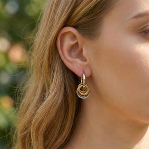 May include: Silver hoop earrings with three interlocking rings, one gold and two silver. The earrings are worn on a person's ear, with the focus on the jewelry. The background is blurred, suggesting an outdoor setting.