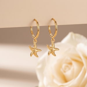 Può includere: Un paio di orecchini a cerchio color oro con ciondoli a forma di stella marina. Gli orecchini sono sospesi dai cerchi e presentano dettagliati disegni di stelle marine. Lo sfondo è beige chiaro con un fiore bianco sfocato.