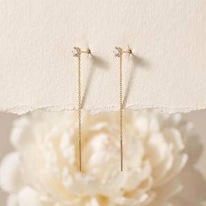 May include: A pair of gold threader earrings, each featuring a single small round clear gemstone set in a six-prong setting at the top, connected to a delicate gold chain that ends in a thin, straight gold bar post for a sleek, dangling look.