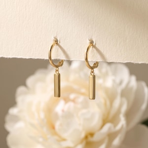 May include: A pair of gold hoop earrings, each featuring a small, smooth, rectangular bar charm suspended from the bottom of the hoop. The earrings are displayed hanging from a textured, off-white paper edge, showcasing a polished metallic finish.