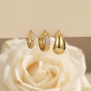 Puede incluir: Tres pendientes de aro dorados de diferentes diseños. Uno es trenzado, otro está incrustado con pequeñas piedras y el tercero tiene forma de lágrima lisa. Se presentan sobre un fondo neutro. Los pendientes son de color dorado cálido.