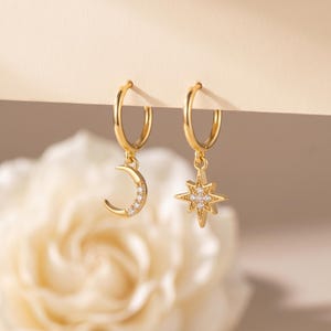 Peut inclure: Une paire de créoles dorées avec des breloques pendantes. Une boucle d'oreille présente un croissant de lune orné de petites pierres claires, et l'autre boucle d'oreille porte une étoile à huit branches sertie de petites pierres claires.