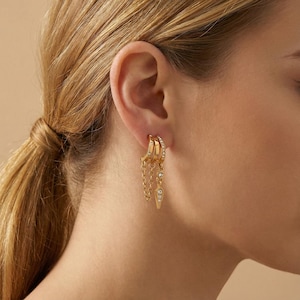 Może przedstawiać: Złote kolczyki o warstwowym designie. Kolczyki posiadają wiele obręczy, niektóre ozdobione małymi, przezroczystymi kryształkami. Delikatne łańcuszki i wisiorki w kształcie łezki z kryształkami zwisają z obręczy, tworząc stylowy i elegancki wygląd.