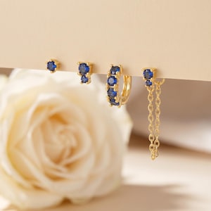 May include: A set of four gold-tone earrings featuring deep blue round-cut stones. The set includes a single stud, a double-stone stud, a small hinged hoop earring with multiple stones, and a double-stone stud connected to a delicate gold chain.