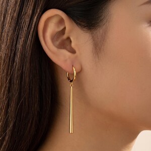 May include: A gold hoop earring featuring a long, smooth, tapered cylindrical drop pendant. The earring is polished with a reflective metallic finish, consisting of a circular hoop attached to a slender, vertical bar that hangs straight down.
