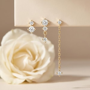 May include: Three pairs of gold-toned earrings with clear, sparkling gemstones. One pair is a stud design, another has three gemstones in a drop style, and the third features a chain with a single dangling gemstone.