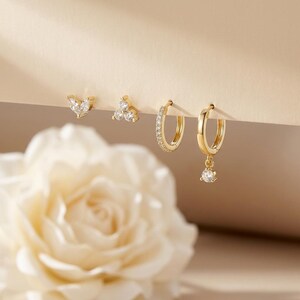 May include: A collection of gold-toned earrings. The set includes two pairs of stud earrings with clear gemstones, and two pairs of hoop earrings, one with a row of gemstones and one with a dangling gemstone. A white rose is in the foreground.