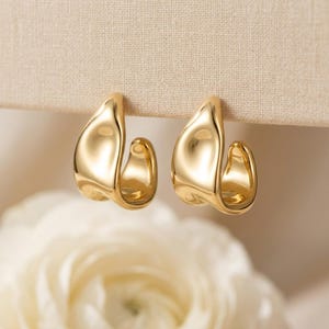 May include: A pair of gold-colored hoop earrings. The earrings have a smooth, curved design with a glossy finish. The earrings are displayed against a neutral background, with a soft, blurred floral element in the lower portion of the image.