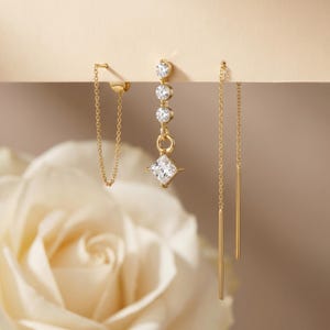 Könnte beinhalten: Eine Sammlung goldener Ohrringe. Ein Ohrring hat eine Kette, die vom Ohr herabhängt. Ein weiterer Ohrring hat drei runde Edelsteine und einen quadratischen Edelstein. Der dritte Ohrring hat zwei goldene Stäbe.