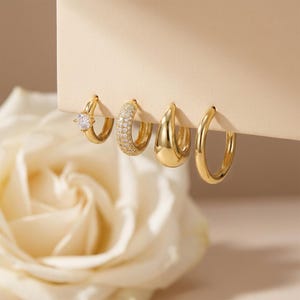 Könnte beinhalten: Vier goldene Creolen. Ein Ohrring hat einen einzelnen klaren Edelstein, ein anderer ist mit kleinen klaren Steinen besetzt, und zwei sind aus massivem Gold. Die Ohrringe werden vor einem neutralen Hintergrund mit einer verschwommenen weißen Rose präsentiert.
