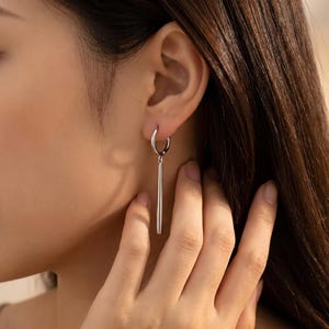 May include: A silver-toned earring featuring a small circular hoop attached to a smooth, cylindrical drop pendant. The metal has a polished, reflective finish, and the design consists of a simple vertical bar suspended from the hoop earring.