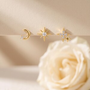 May include: Three gold-tone stud earrings featuring celestial motifs. One earring is a crescent moon with a single clear stone, one is an eight-pointed star with a central clear stone, and one is a four-pointed star cluster with multiple clear stones.