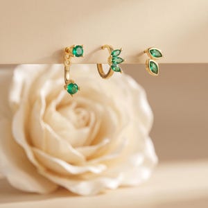 May include: Three gold-tone earrings featuring green gemstones. The first is a curved bar with two round green stones, the second is a small hoop with three marquise-cut green stones, and the third is a stud with two marquise-cut green stones.