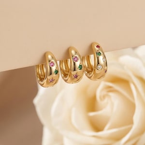 May include: Three gold-colored hoop earrings with colorful gemstones. Each earring features a curved design and is adorned with pink, green, and clear gemstones. The earrings are displayed against a soft, neutral background, with a white rose in the background.