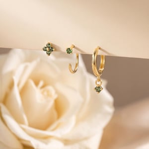 May include: Three gold earrings featuring green gemstones. One earring is a four-stone flower stud, one is a small open hoop with a single green stone, and one is a gold hoop earring with a dangling green gemstone charm attached to the bottom.