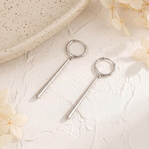 May include: A pair of silver-toned hoop earrings, each featuring a smooth, cylindrical bar pendant suspended from the circular hoop. The earrings have a polished, reflective metallic finish and a simple, clean design with no additional embellishments.