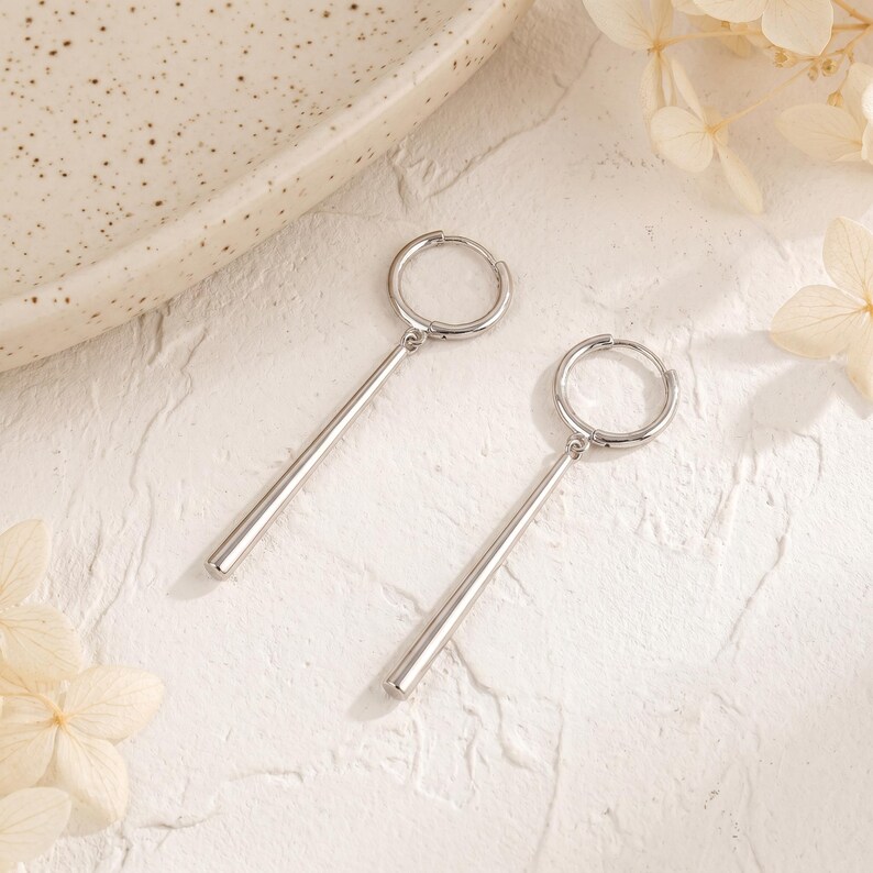 May include: A pair of silver-toned hoop earrings, each featuring a smooth, cylindrical bar pendant suspended from the circular hoop. The earrings have a polished, reflective metallic finish and a simple, clean design with no additional embellishments.