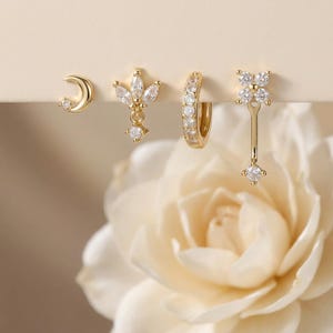 May include: A collection of four gold-toned earrings. The earrings include a crescent moon stud, a floral stud with a dangling charm, a hoop earring, and a drop earring with a four-petal flower design and a dangling charm.