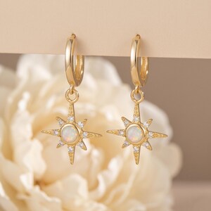 May include: A pair of gold-colored hoop earrings. Each earring features a starburst pendant with a central opal-like stone and surrounding clear crystals. The earrings are displayed against a soft, neutral background.