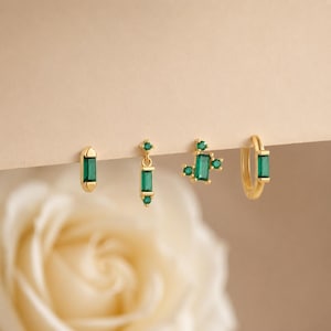 May include: A set of four gold earrings featuring emerald green baguette-cut stones. The collection includes a simple baguette stud, a dangling baguette charm, a cluster of three baguette stones, and a small gold hoop with a baguette stone.