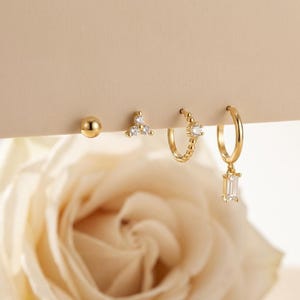 May include: A collection of gold earrings, including a ball stud, a triangle stud with three clear stones, a small hoop with a single clear stone, and a larger hoop with a rectangular clear stone pendant. The earrings are displayed against a neutral background.