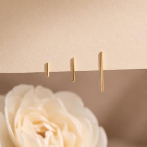 May include: Three gold-toned metal bar earrings of varying lengths arranged in a row. Each earring consists of a thin, straight, vertical rod with a smooth, polished finish. The earrings are displayed against a plain, light-colored surface.